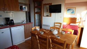Een compacte keuken met een magnetron, koffiezetapparaat en vaatwasser. Een eettafel met stoelen staat in het midden, en een bed met een rode sprei op de achtergrond.