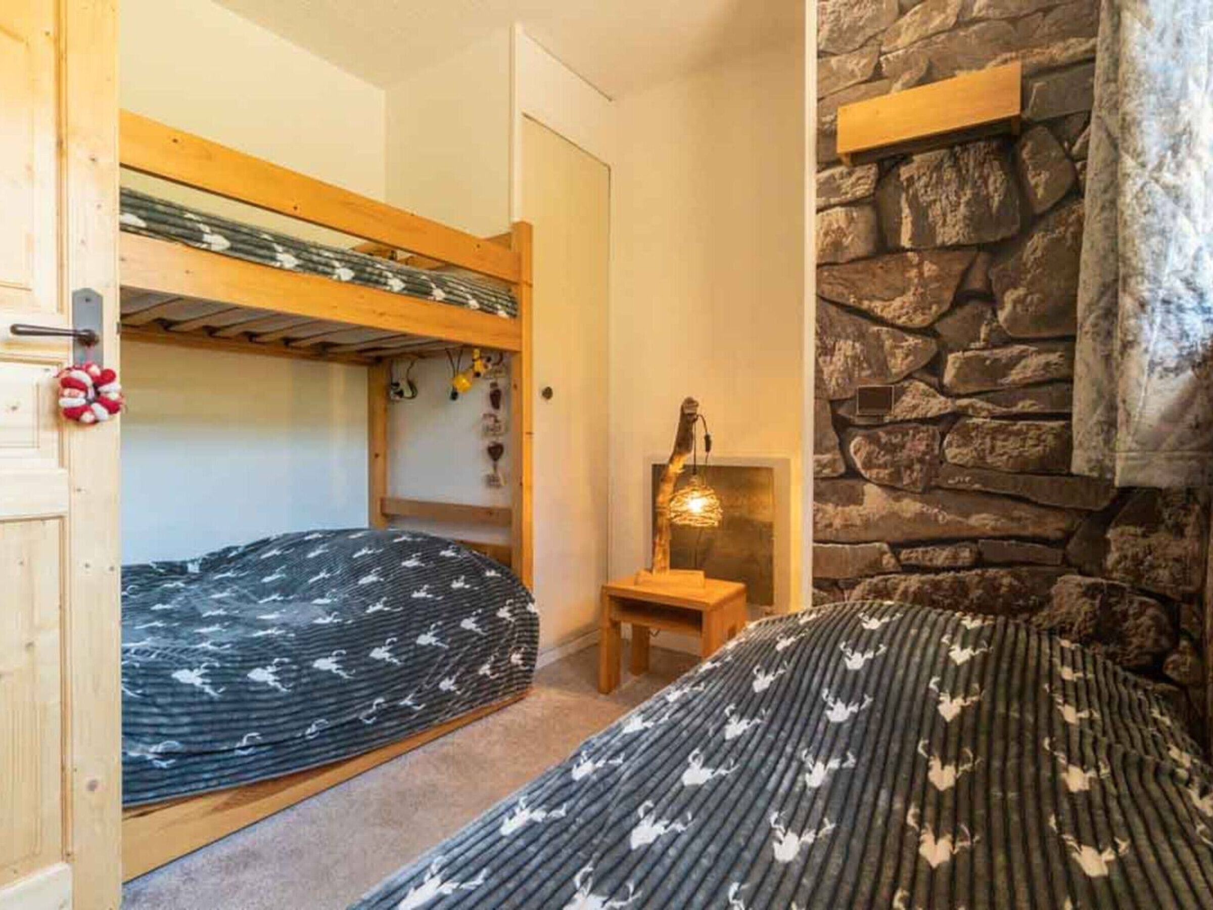 A room with bunk beds, a stone wall, a wooden shelf, and a small table with a lamp.