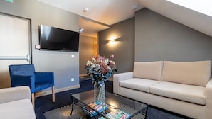A modern living room with a beige sofa, a blue armchair, a glass coffee table with magazines, a flat-screen TV mounted on the wall, and a decorative vase with flowers. - Madame Vacances Hôtel les Cimes (Vaujany)