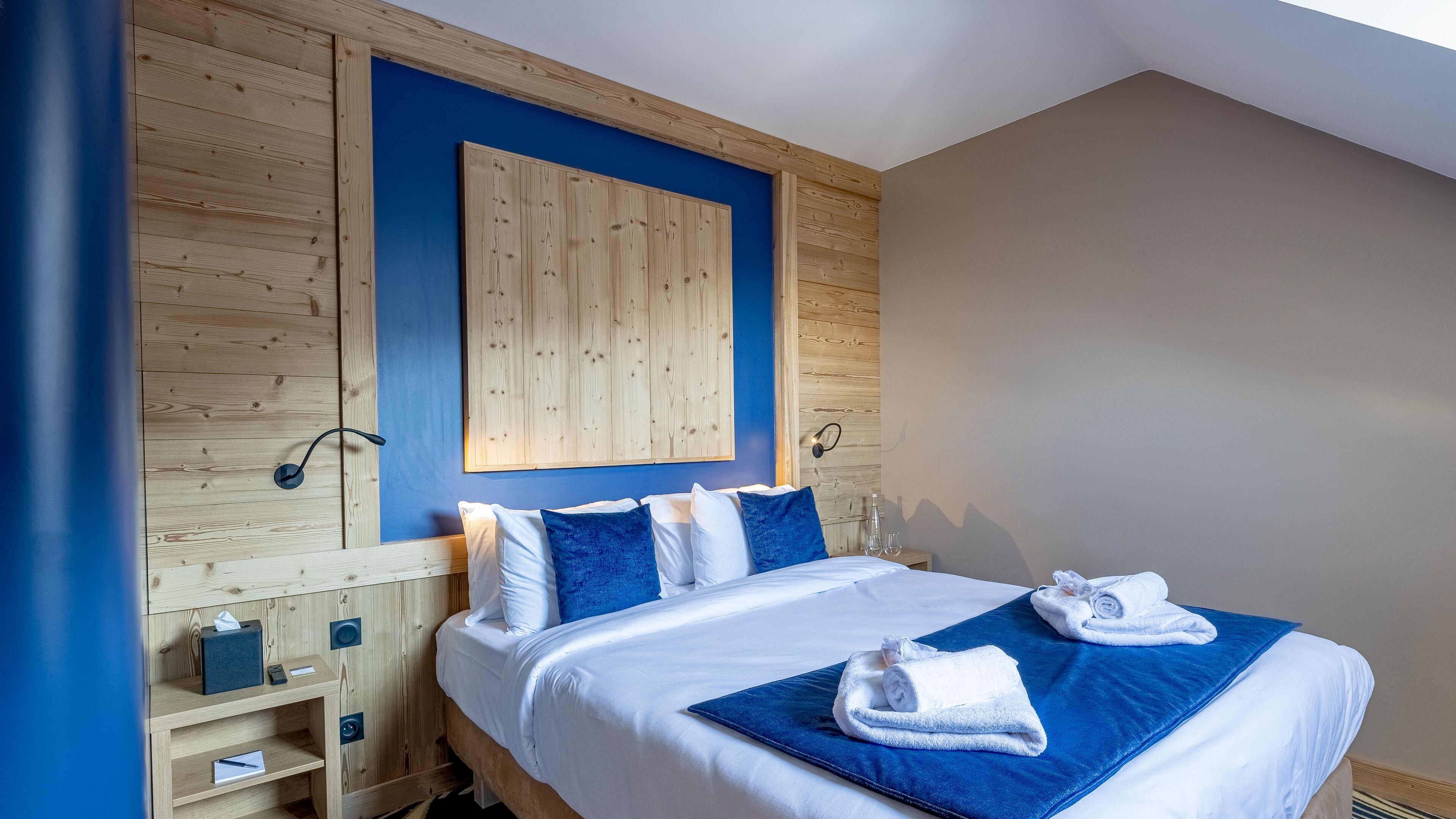 Ett hotellrum med en sänggavel i trä, en stor säng med vita sängkläder och blå kuddar, ett nattduksbord med en lampa och ett konstverk som hänger på väggen.