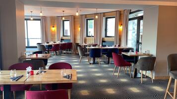 Ein Restaurant mit mehreren Tischen und Stühlen, Holzdielen und großen Fenstern mit Vorhängen.