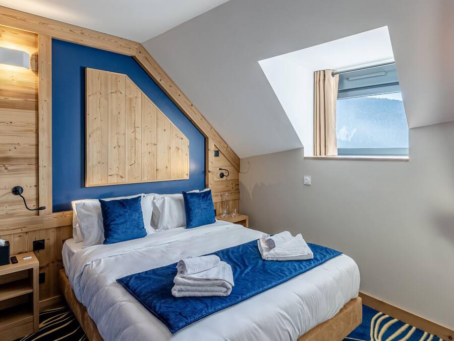 A bedroom with a sloped ceiling, a skylight, a bed with white bedding and blue pillows, a wooden headboard, and a small bedside table with a lamp.
