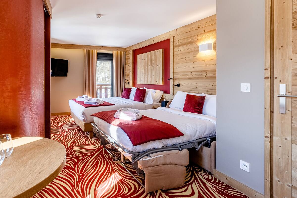 Ein Hotelzimmer mit zwei Betten, einem hölzernen Kopfteil, einem Teppich mit roten und weißen Mustern, einem kleinen runden Tisch mit Gläsern und einem an der Wand montierten Fernseher.