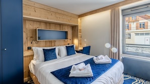 A hotel room with a wooden wall panel, a bed with white and blue bedding, a blue throw blanket, and a window with a view of buildings. - Madame Vacances Hôtel les Cimes (Vaujany)