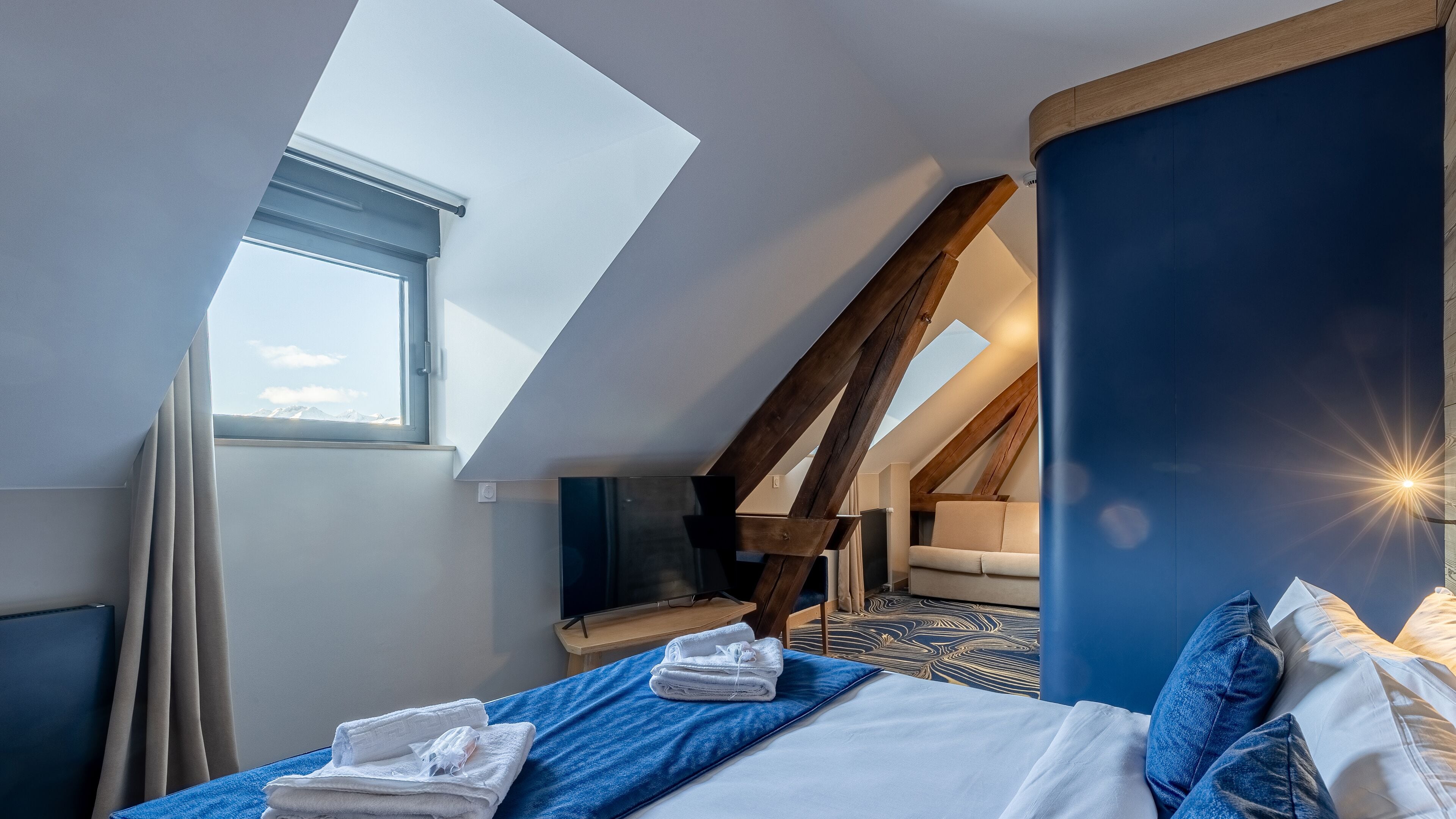 Quarto com claraboia, cama com roupa de cama azul, teto de madeira e vista para o céu.