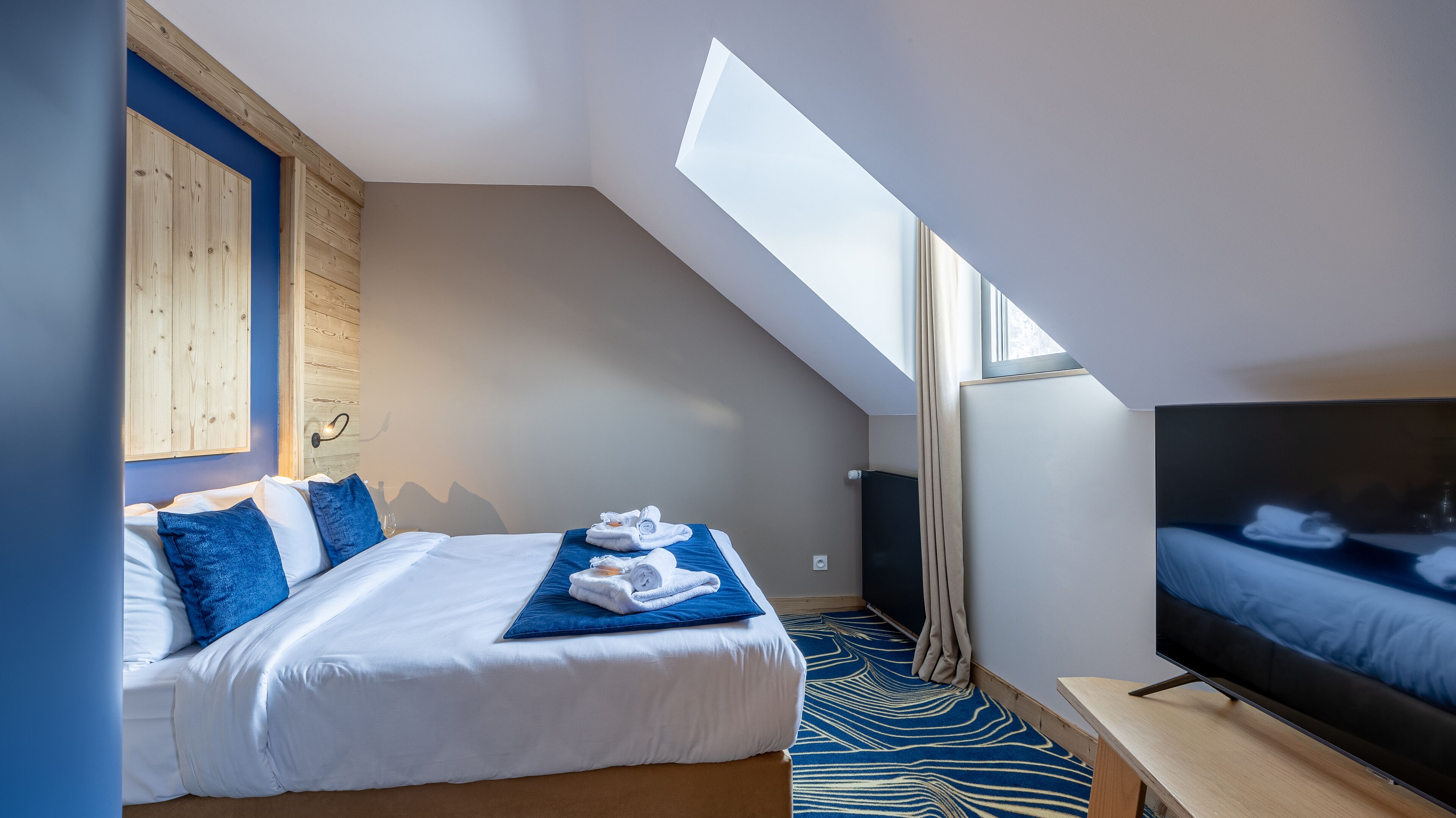 A modern hotel room with a bed, a TV, a wooden accent wall, and a skylight.