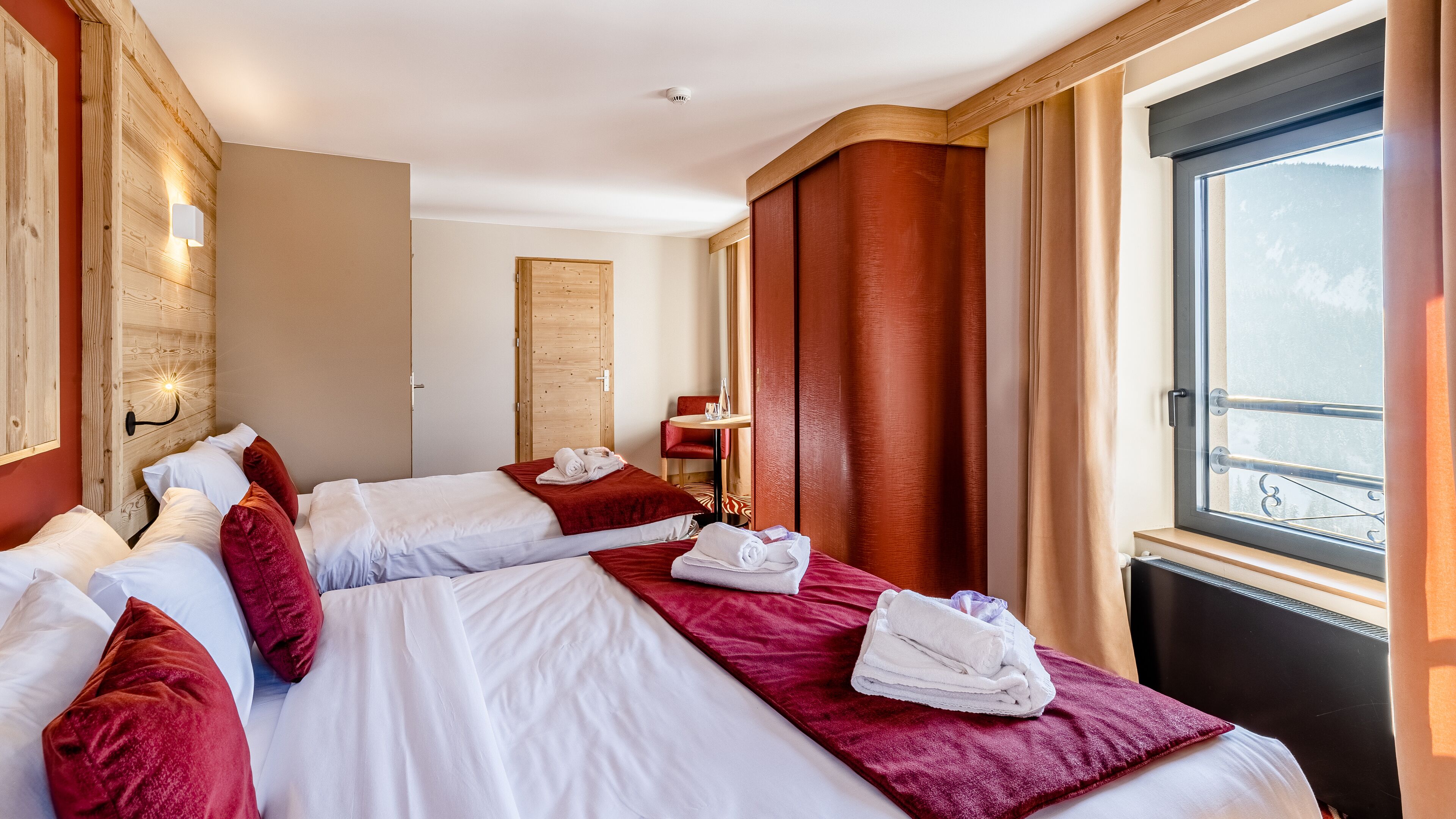 Habitación de hotel con dos camas, cabecero de madera, ventana con vistas y una pequeña mesa con una silla.