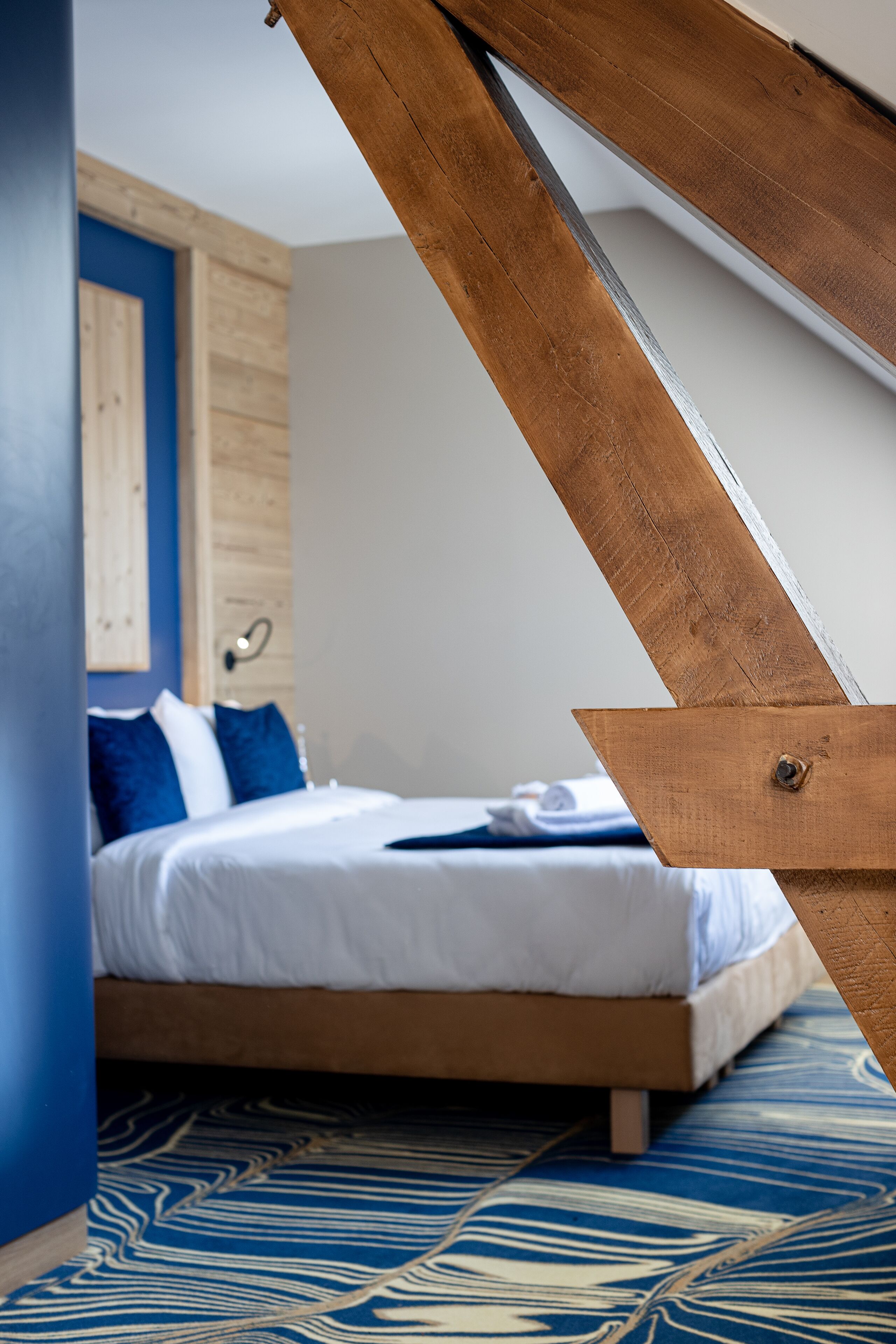 Un dormitorio con cama, ropa de cama azul y blanca, cabecero de madera y alfombra estampada.