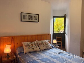 Une chambre à coucher avec une tête de lit en bois, un lit recouvert d’une literie à motifs, une lampe de chevet et une fenêtre donnant sur des arbres.