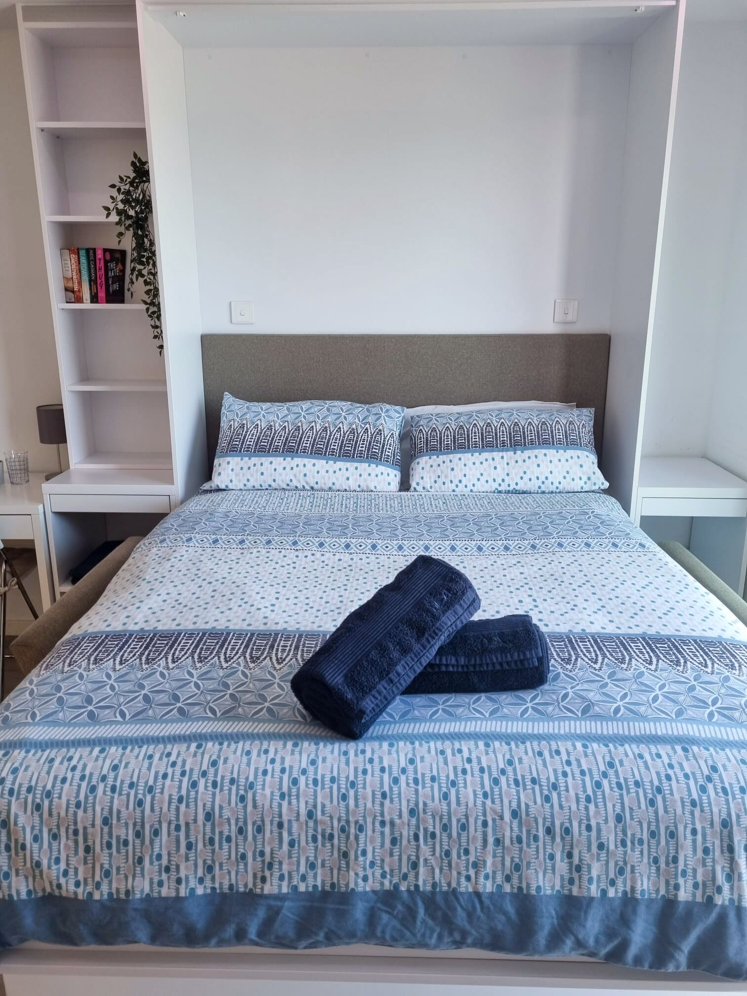 Un lit bien fait, avec une literie à motifs, une table de chevet avec une lampe et une bibliothèque remplie de livres et agrémentée d’une plante.