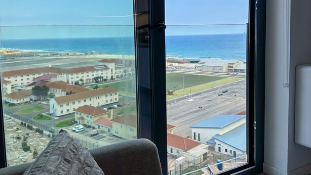 A room with a large window overlooking a coastal area, a bed, and a view of buildings and a road.