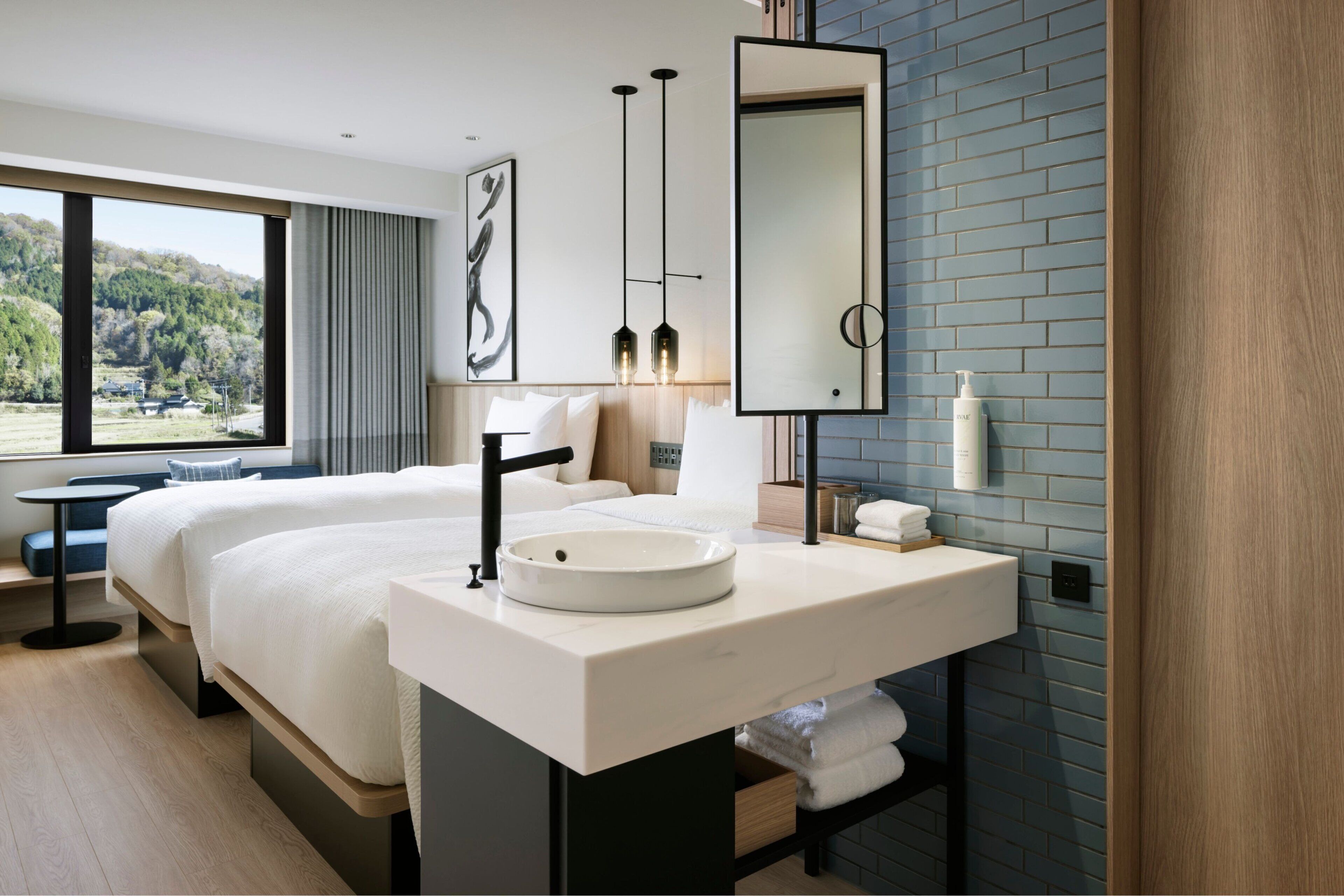 A modern hotel room with a large bed, a round sink, a mirror, and a view of a forest through large windows.
