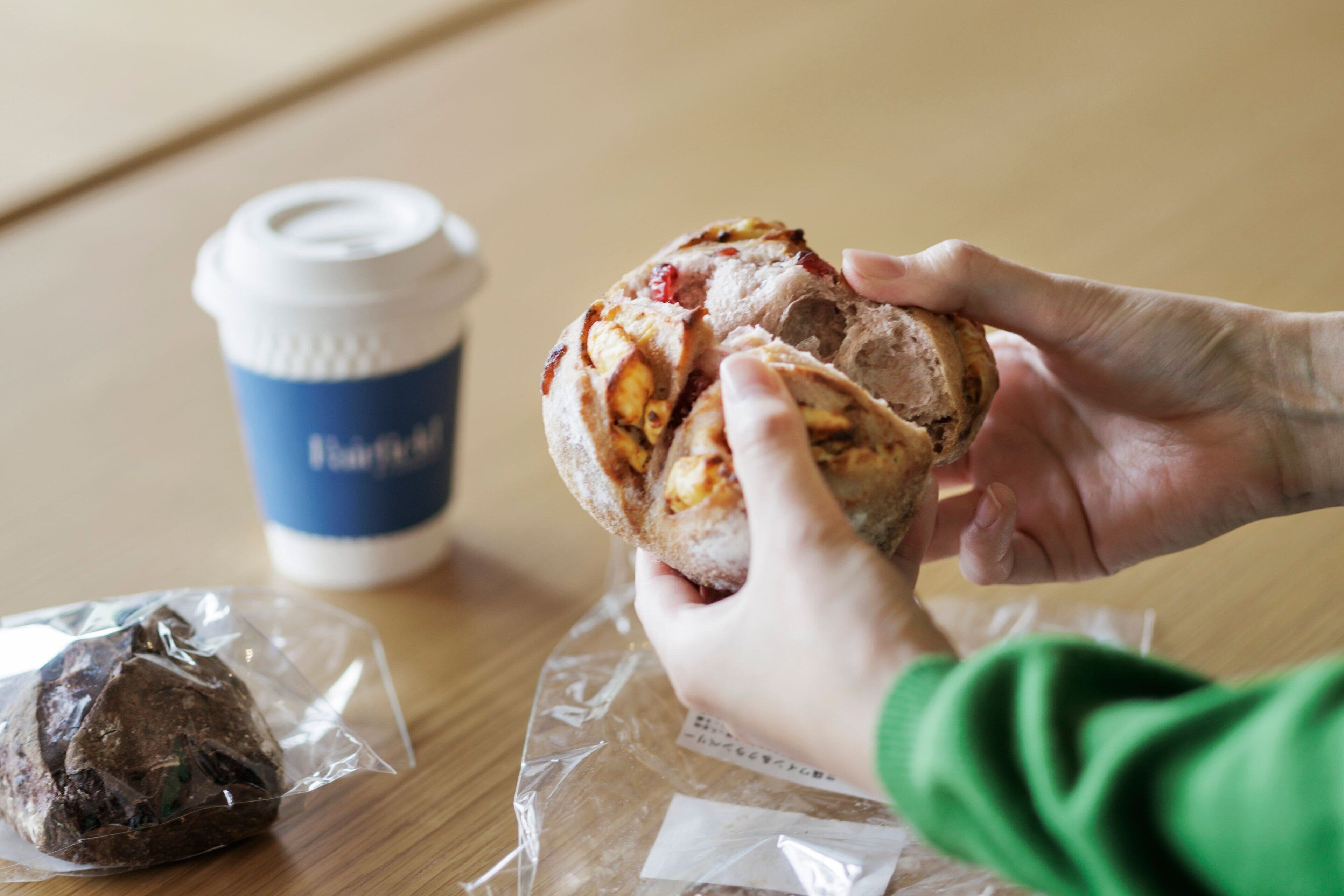 A person holding a pastry with a cup of coffee and a wrapped item on a table.