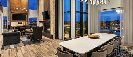 A modern living room with a fireplace, a large dining table, and a view of the city at dusk.