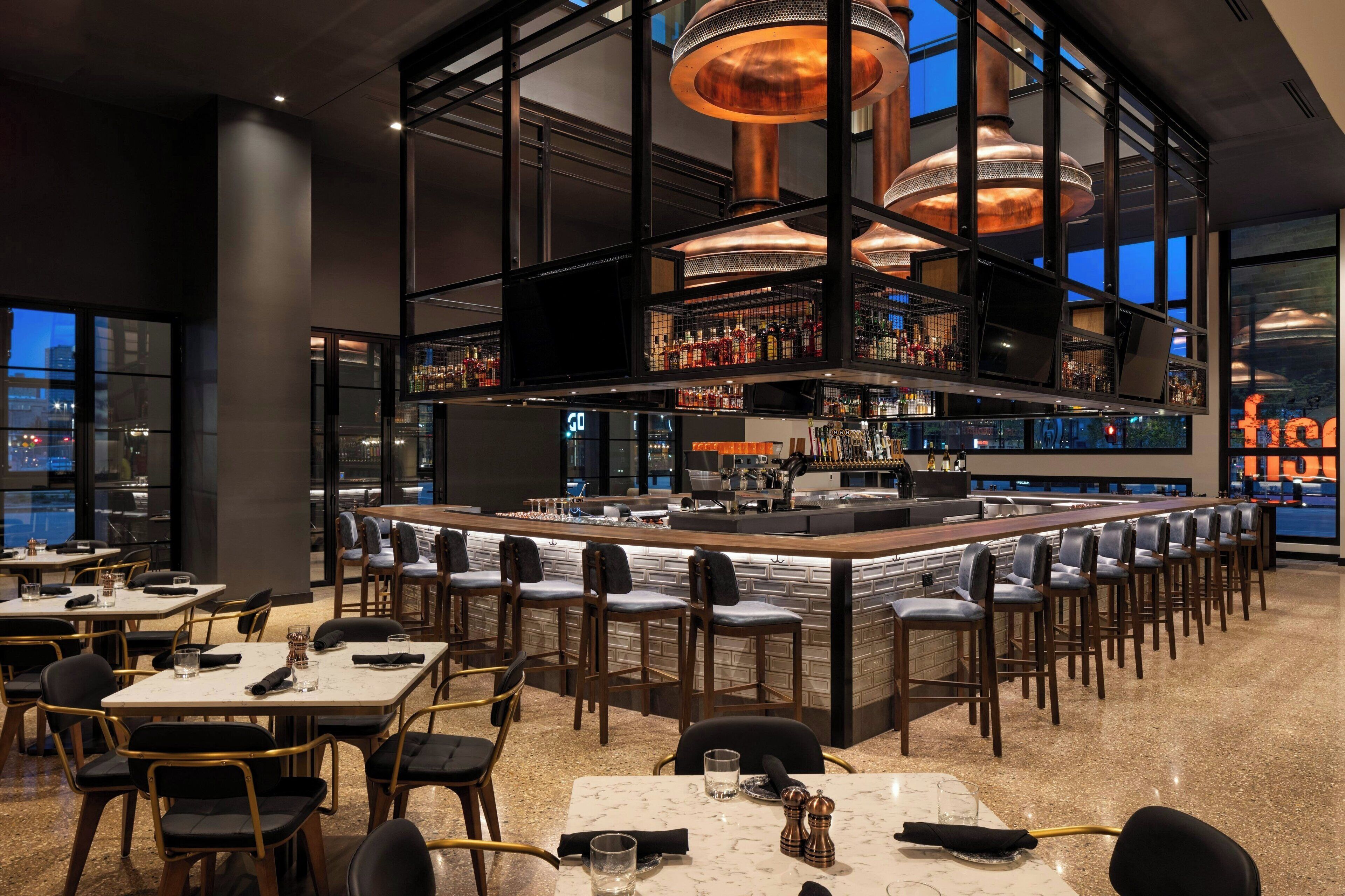 A modern bar area with high ceilings, copper lighting fixtures, and a bar counter with stools.