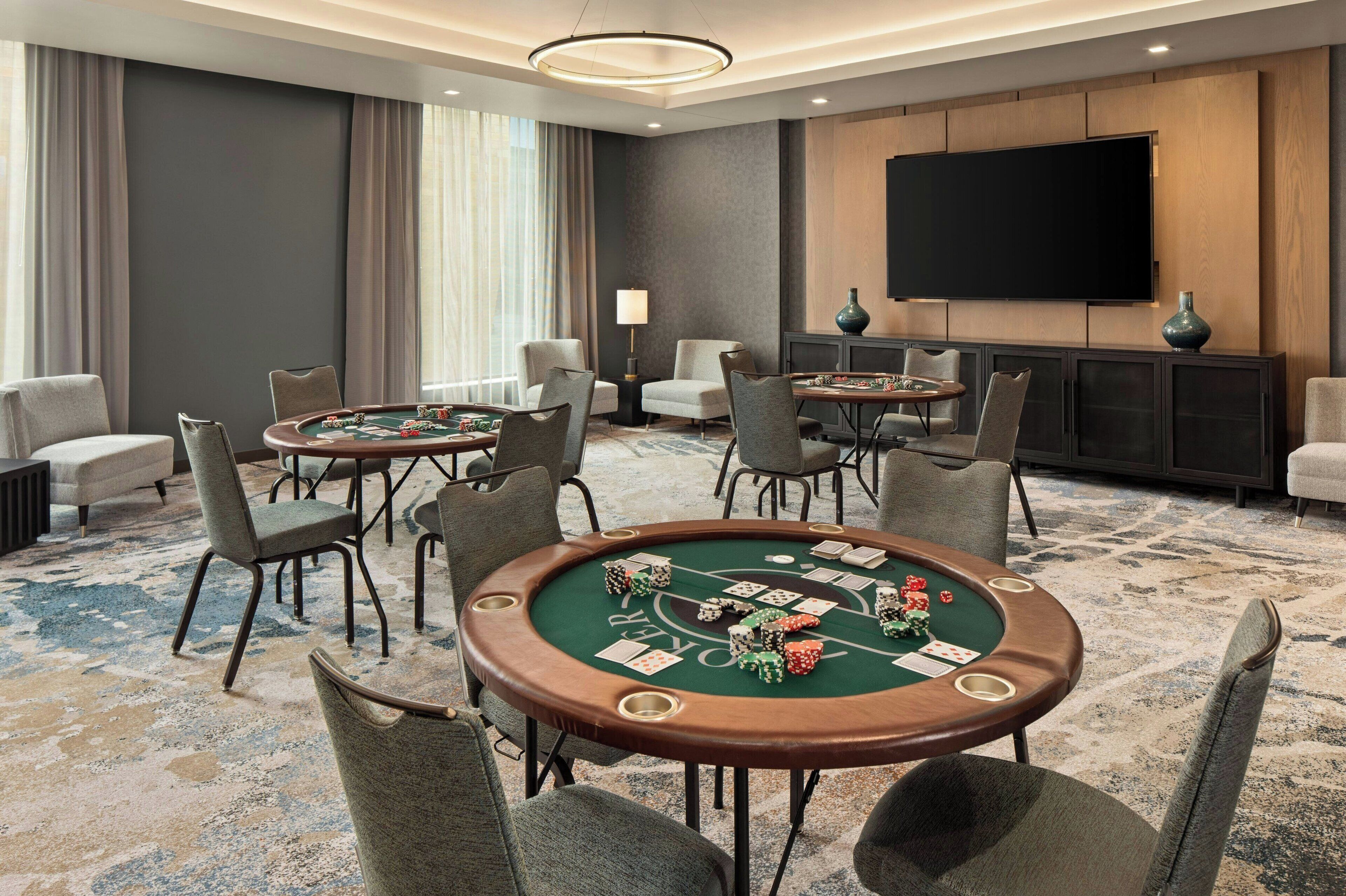 A room with a green felt blackjack table surrounded by chairs, a large flat-screen TV on the wall, and a sofa.