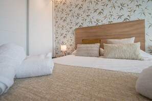 Ein ordentlich bezogenes Bett mit weißen Leinen und einer beige farbenen Tagesdecke, ein Holz-Kopfteil und eine Wand mit Bambusblatt-Tapete.