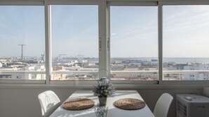 A dining area with a table set for two, a vase with flowers, and a view of a cityscape through large windows. - Apartment La Atalaya de Cádiz (Cádiz)