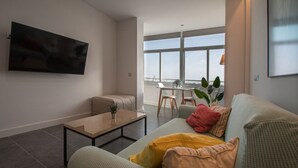 A modern living room with a sofa, a coffee table, and a TV mounted on the wall. There is a dining area with a table and chairs visible through large windows. - Apartment La Atalaya de Cádiz (Cádiz)