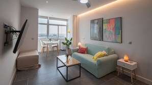 A modern living room with a sofa, a coffee table, and a side table with a lamp. There is a dining area with a table and chairs visible through a large window. - Apartment La Atalaya de Cádiz (Cádiz)