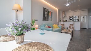 A modern living room with a white sofa, a round coffee table, and a dining table. There is a kitchen area with white cabinets and a ceiling fan. - Apartment La Atalaya de Cádiz (Cádiz)