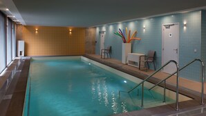 An indoor swimming pool with clear water, a tiled wall, and a seating area with chairs and a table.