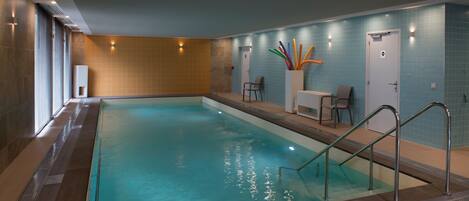 An indoor swimming pool with clear water, a tiled wall, and a seating area with chairs and a table.