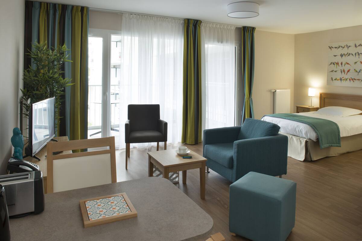 Une chambre d’hôtel avec un lit, un canapé, une chaise, une petite table, une télévision et une fenêtre avec des rideaux.