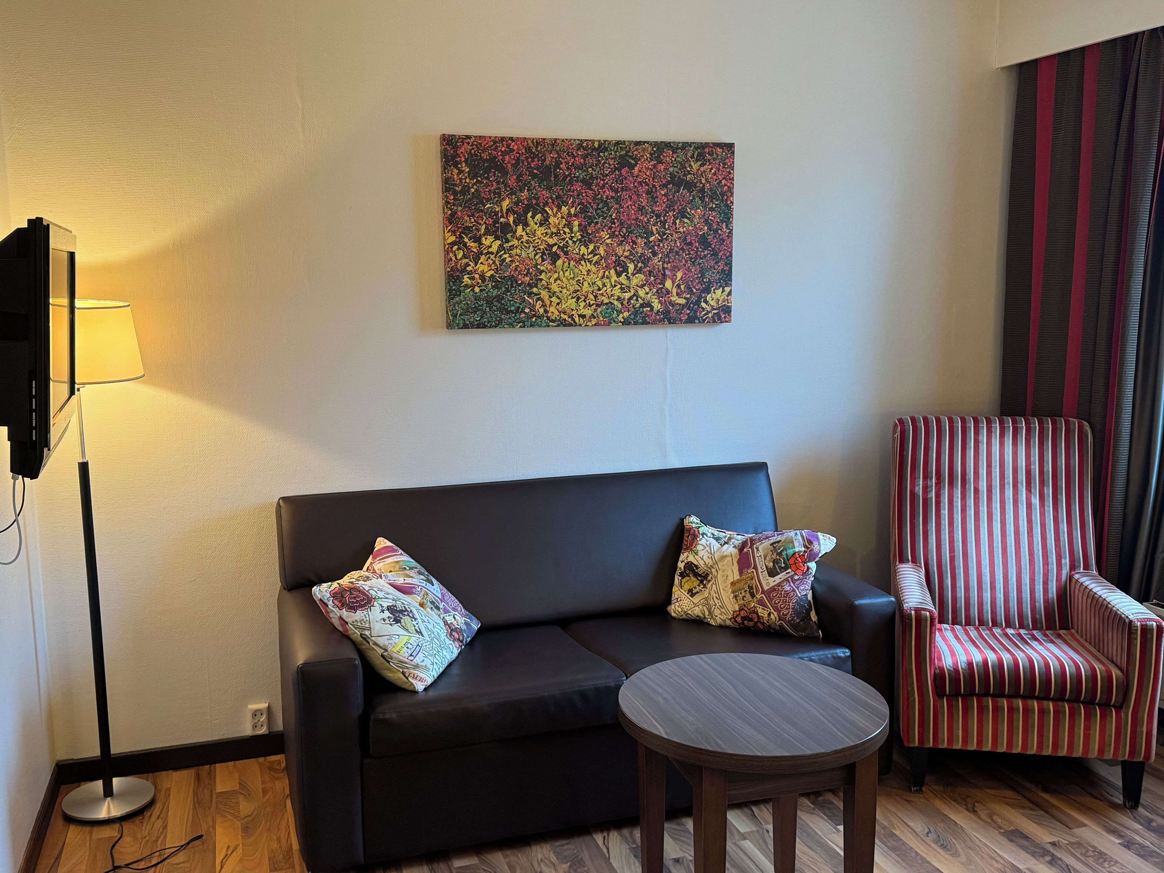 A living room with a black sofa, two patterned pillows, a striped armchair, a round wooden table, and a colorful abstract painting on the wall.