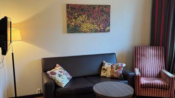 A living room with a black sofa, two patterned pillows, a striped armchair, a round wooden table, and a colorful abstract painting on the wall.