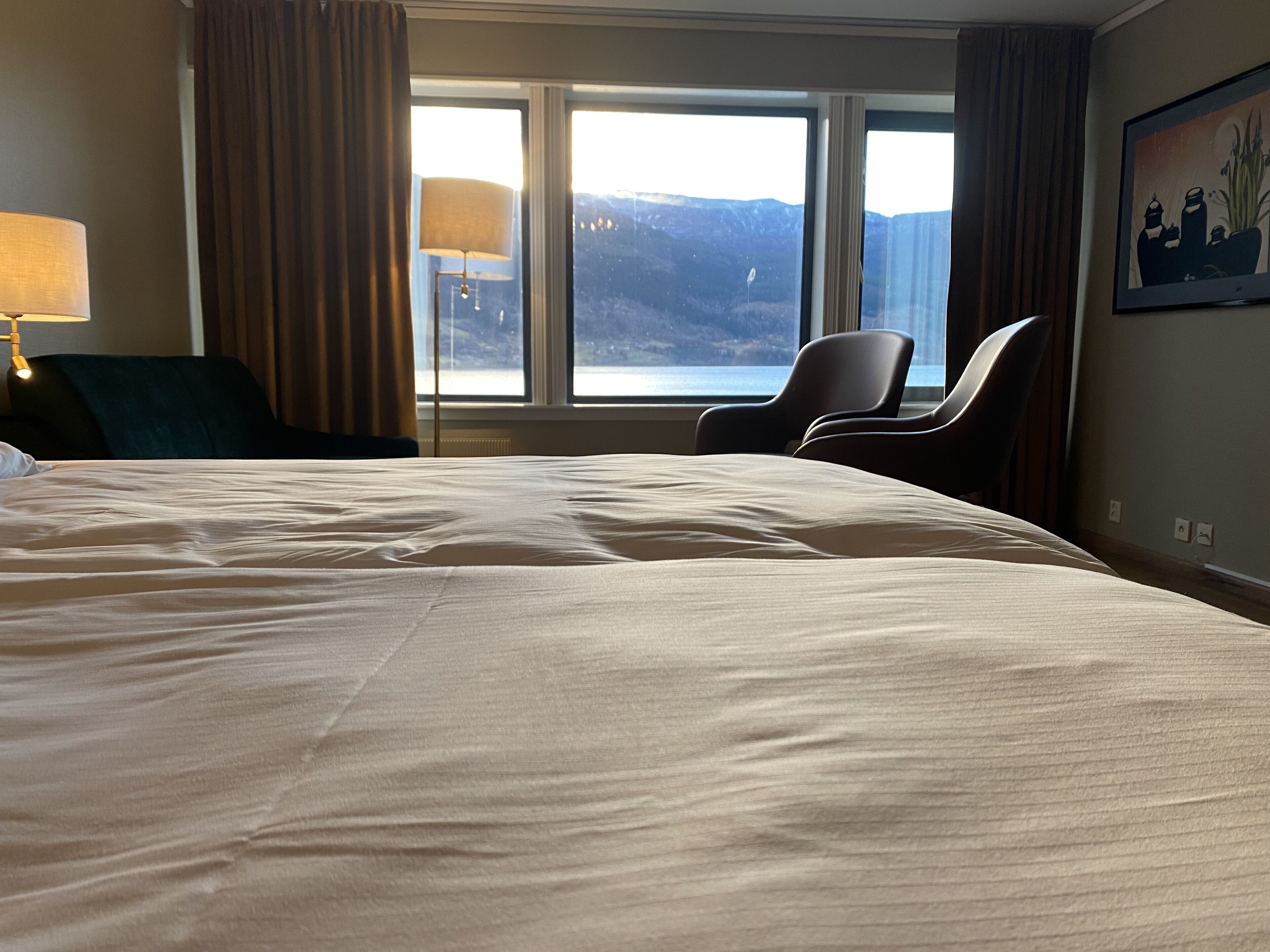 Habitación de hotel con una cama grande, dos sillones, un escritorio, una lámpara y una ventana con vistas a la montaña.
