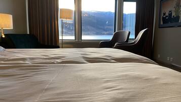 Habitación de hotel con una cama grande, dos sillones, un escritorio, una lámpara y una ventana con vistas a la montaña.