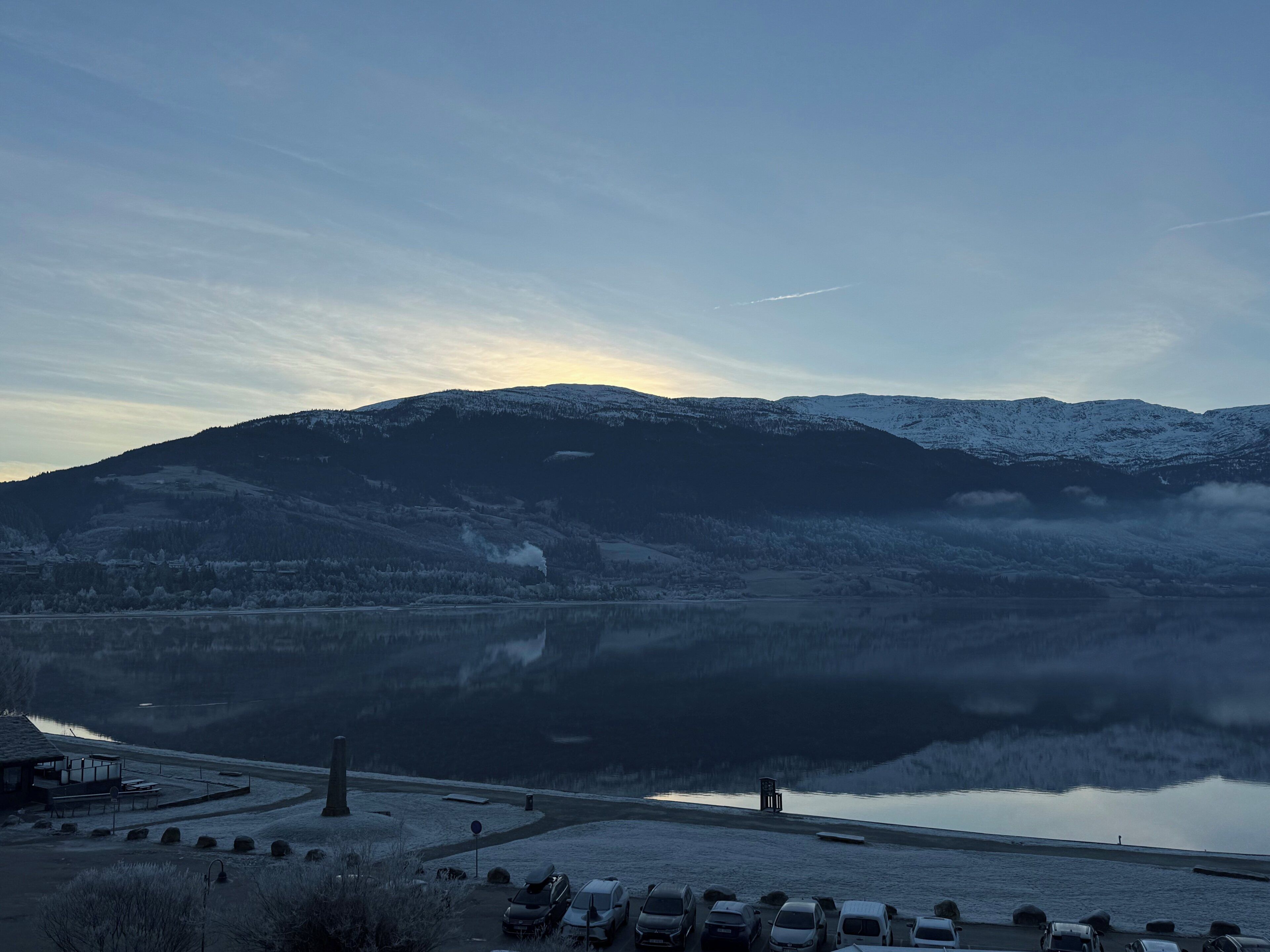 Una vista tranquila de un lago con una montaña nevada al fondo, un estacionamiento con varios coches y un pequeño edificio con techo.