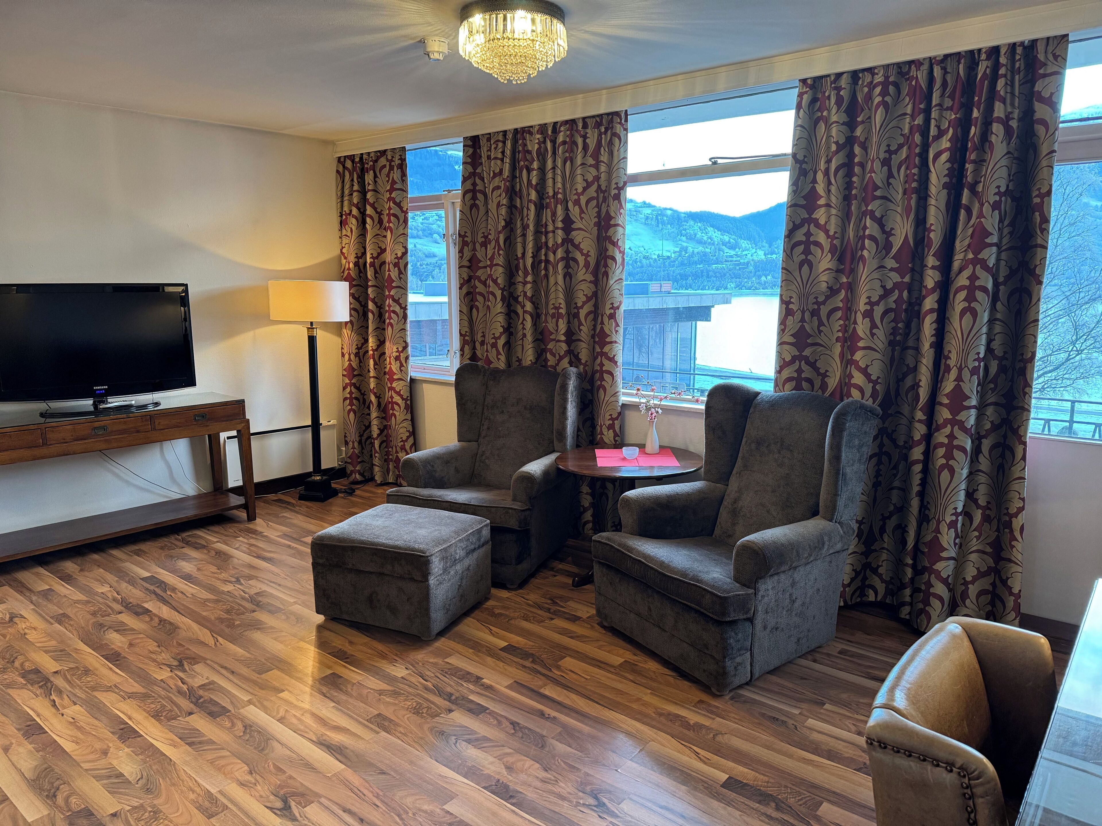 Habitación de hotel con televisión de pantalla plana, dos sillones, una mesita con un jarrón y un ventanal con cortinas.