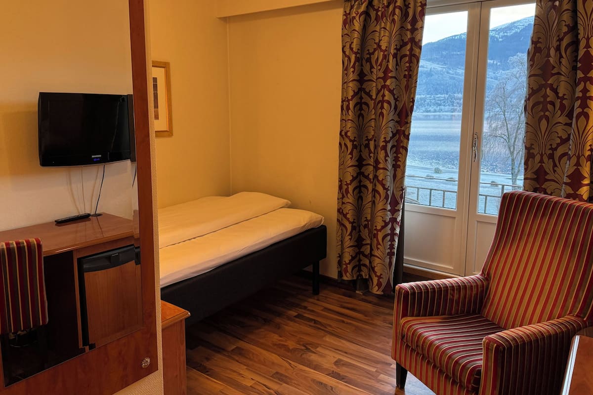Habitació amb un llit, un televisor fixat a la paret, un escriptori de fusta, una butaca rayada i una finestra amb cortines.
