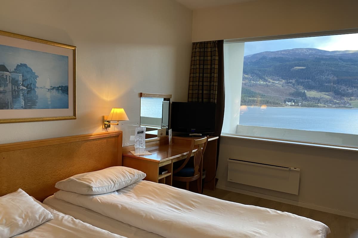 Habitació d'hotel amb dos llits, una taula de nit amb televisió, una làmpada i una finestra que ofereix vistes a un llac i muntanyes.