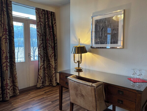 Una habitación con un escritorio de madera, una silla, una lámpara, un cuadro en la pared y una ventana con cortinas.