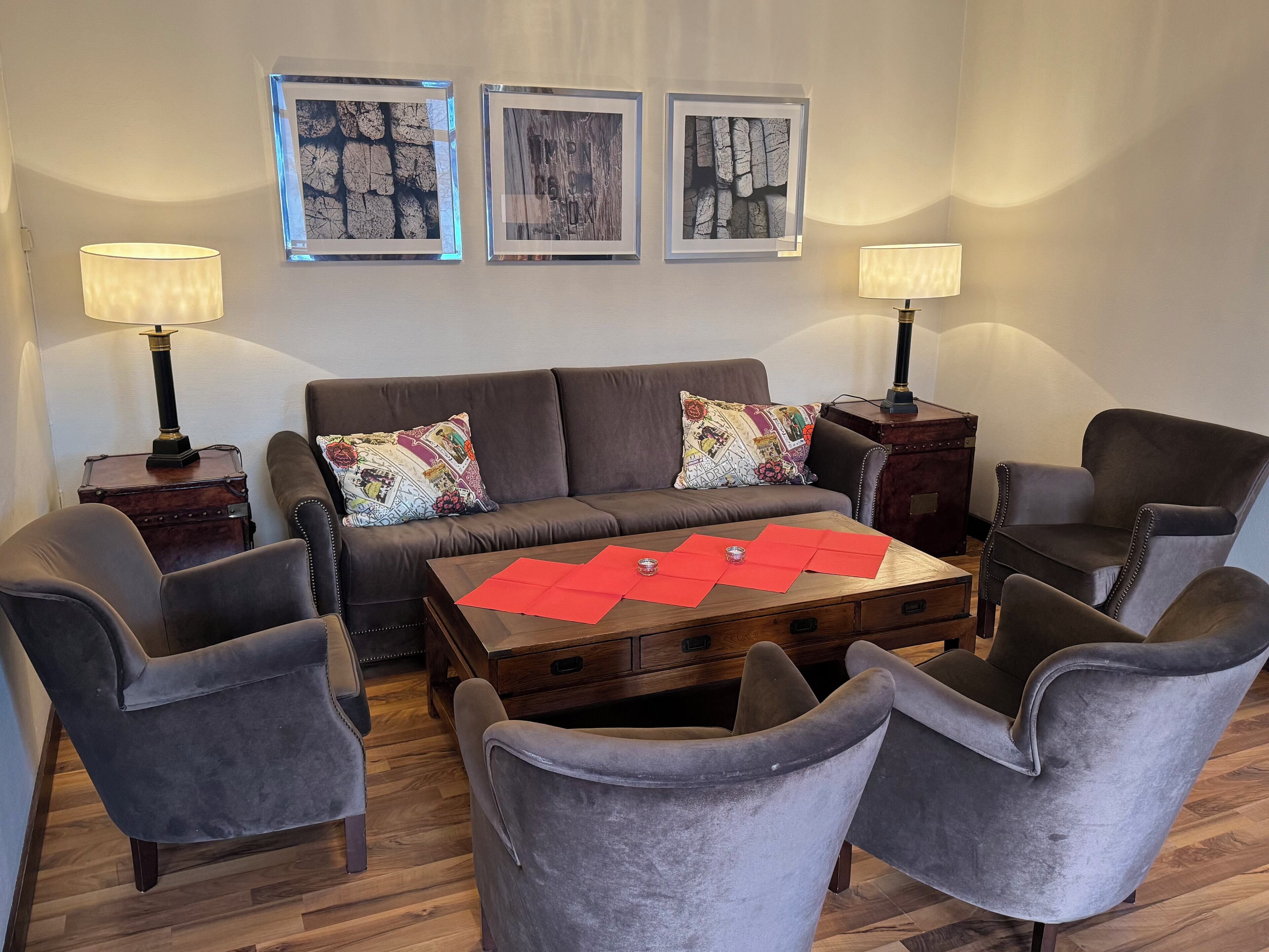 A living room with a brown sofa, two grey armchairs, a wooden coffee table with red placemats, and two wall-mounted lamps.