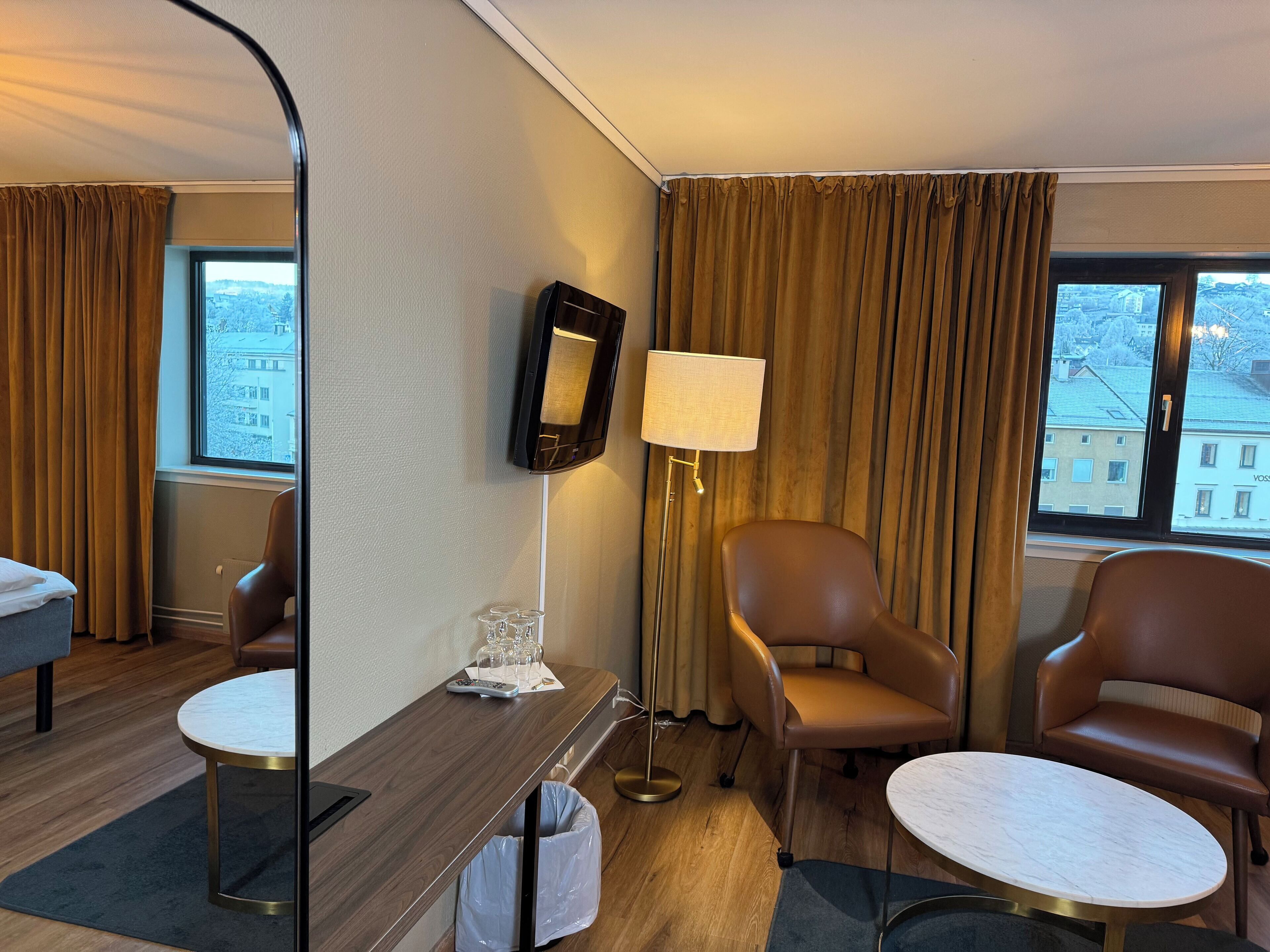 A hotel room with a wooden desk, two chairs, a small table, a mirror, a lamp, and a view of buildings through the window.