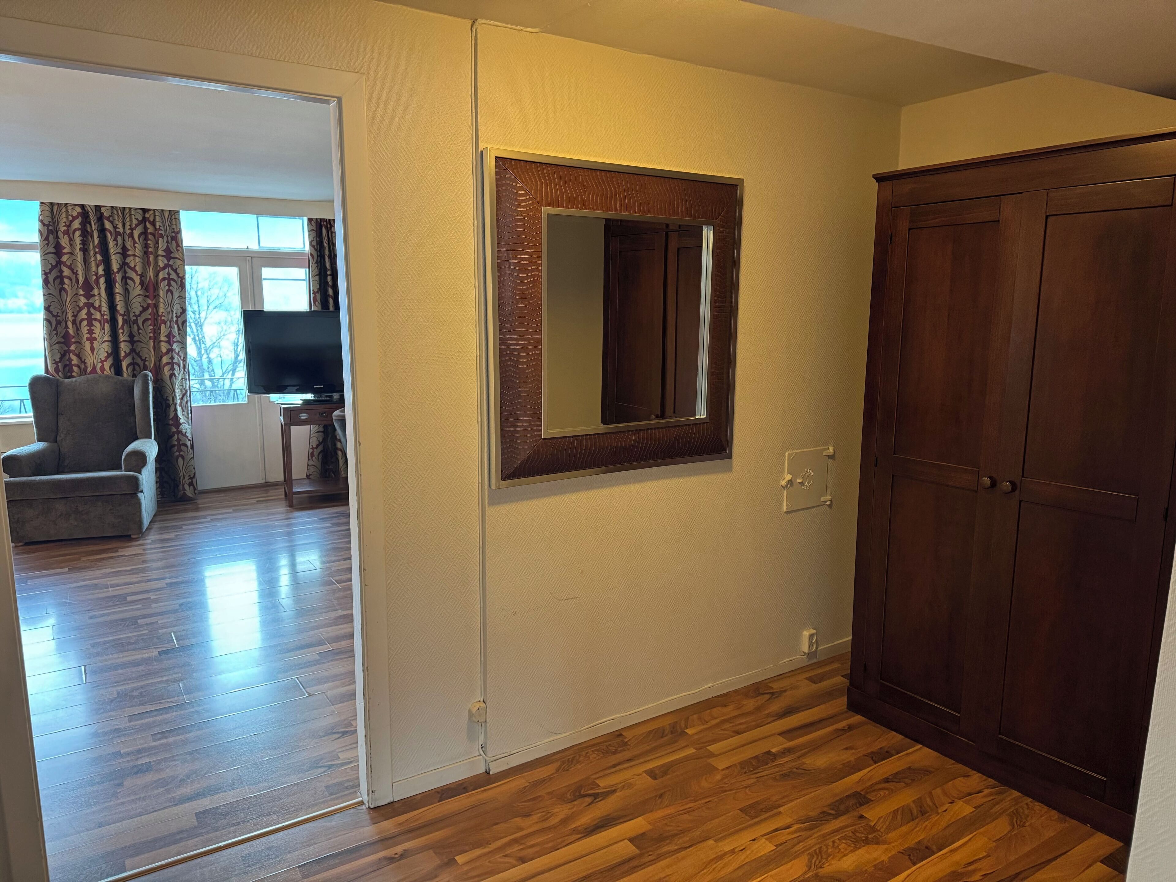 A room with wooden flooring, a large mirror, a wardrobe, and a chair.
