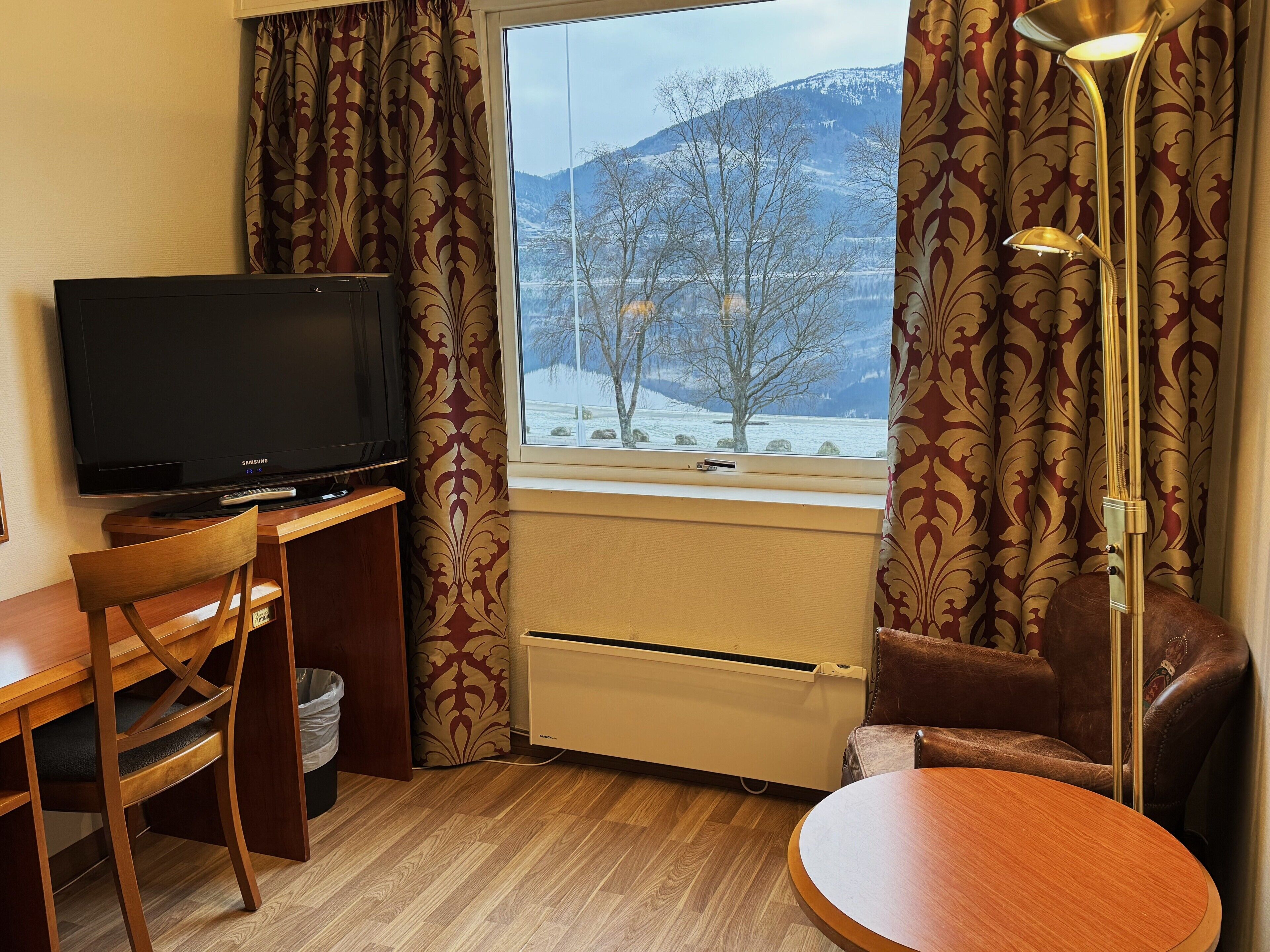 A hotel room with a TV, a desk, a chair, a table, a heater, and a window with a view of snow-covered mountains.