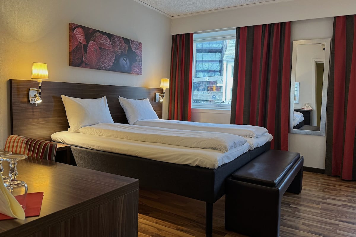 Habitació d'hotel amb un llit de matrimoni, lampades de nit, un panell de capçal de fusta, una panxa, un escriptori amb un got i una finestra amb cortines.