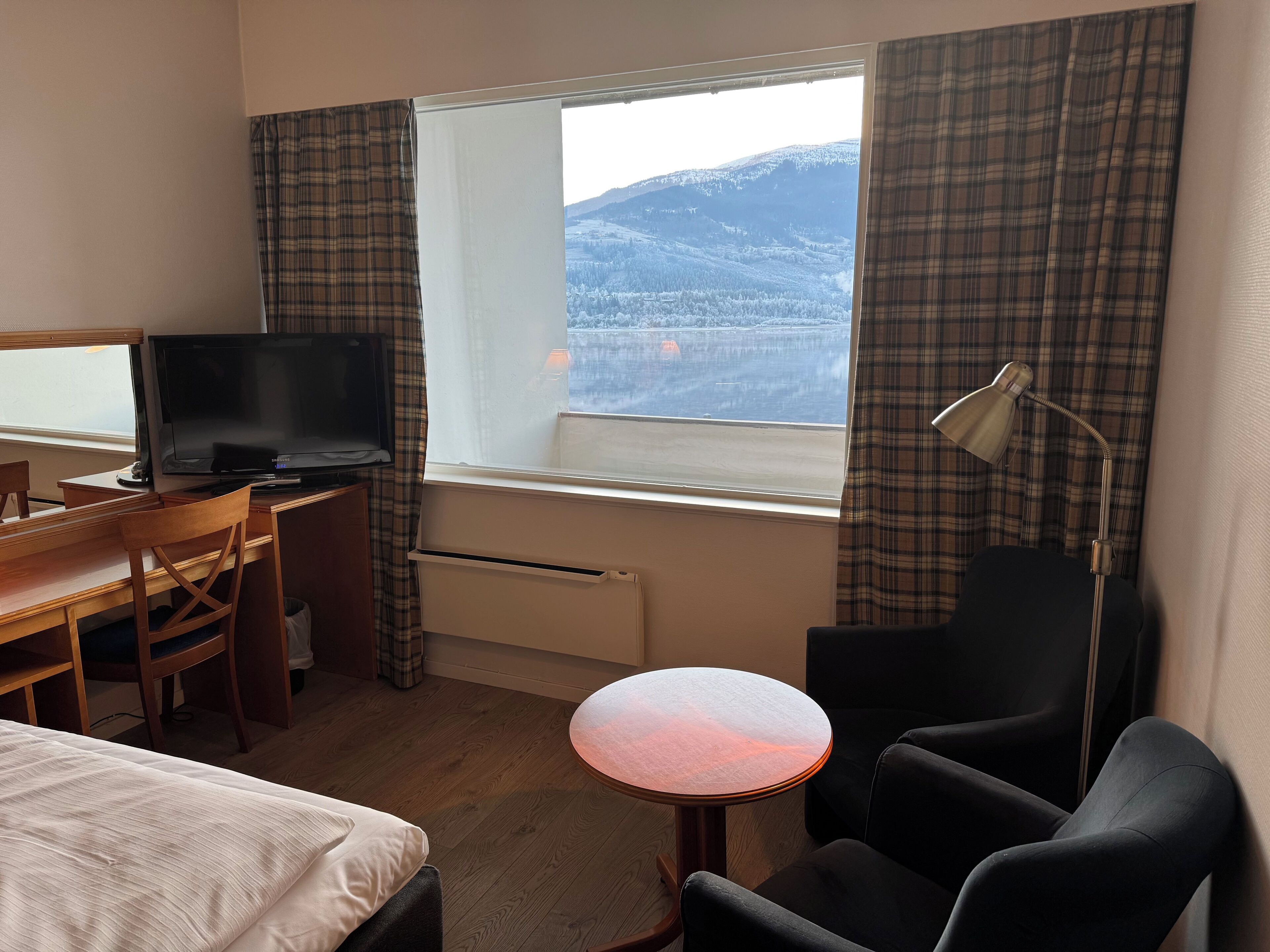 A hotel room with a bed, a desk with a television, a chair, a small table, and a window with a view of a snowy landscape.