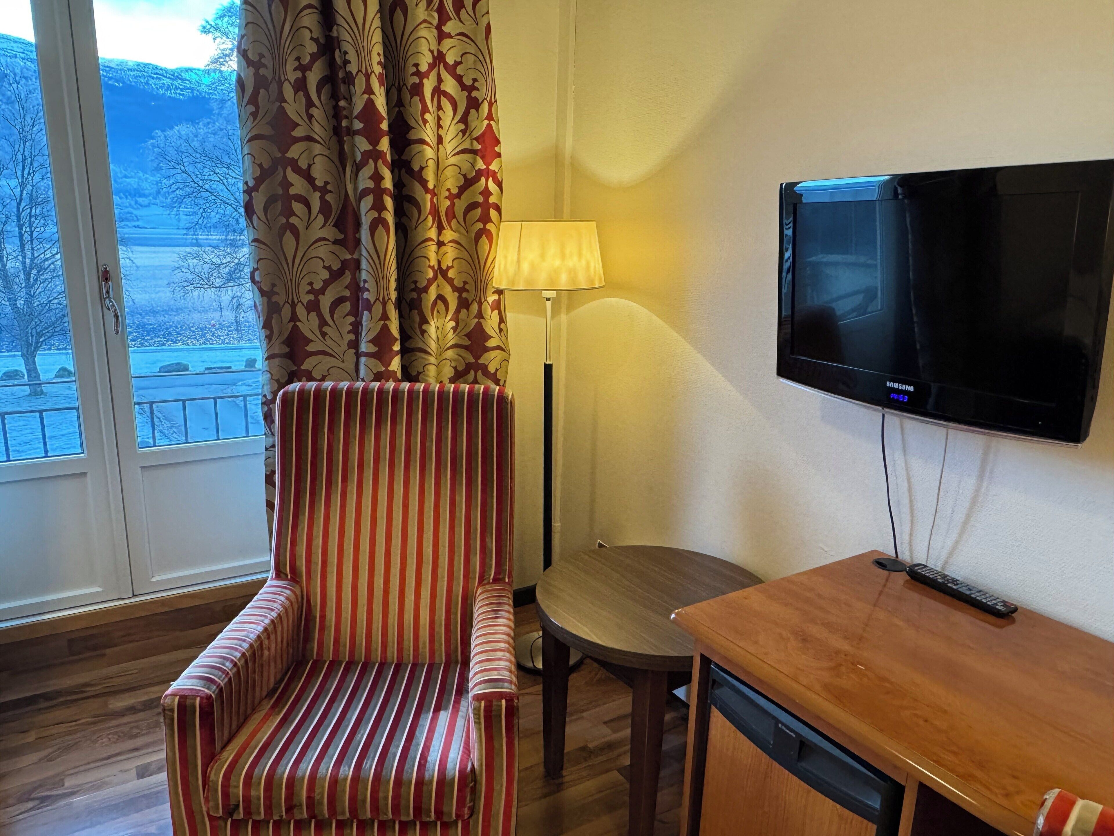 A hotel room with a striped armchair, a small wooden table, a lamp, and a mounted TV.