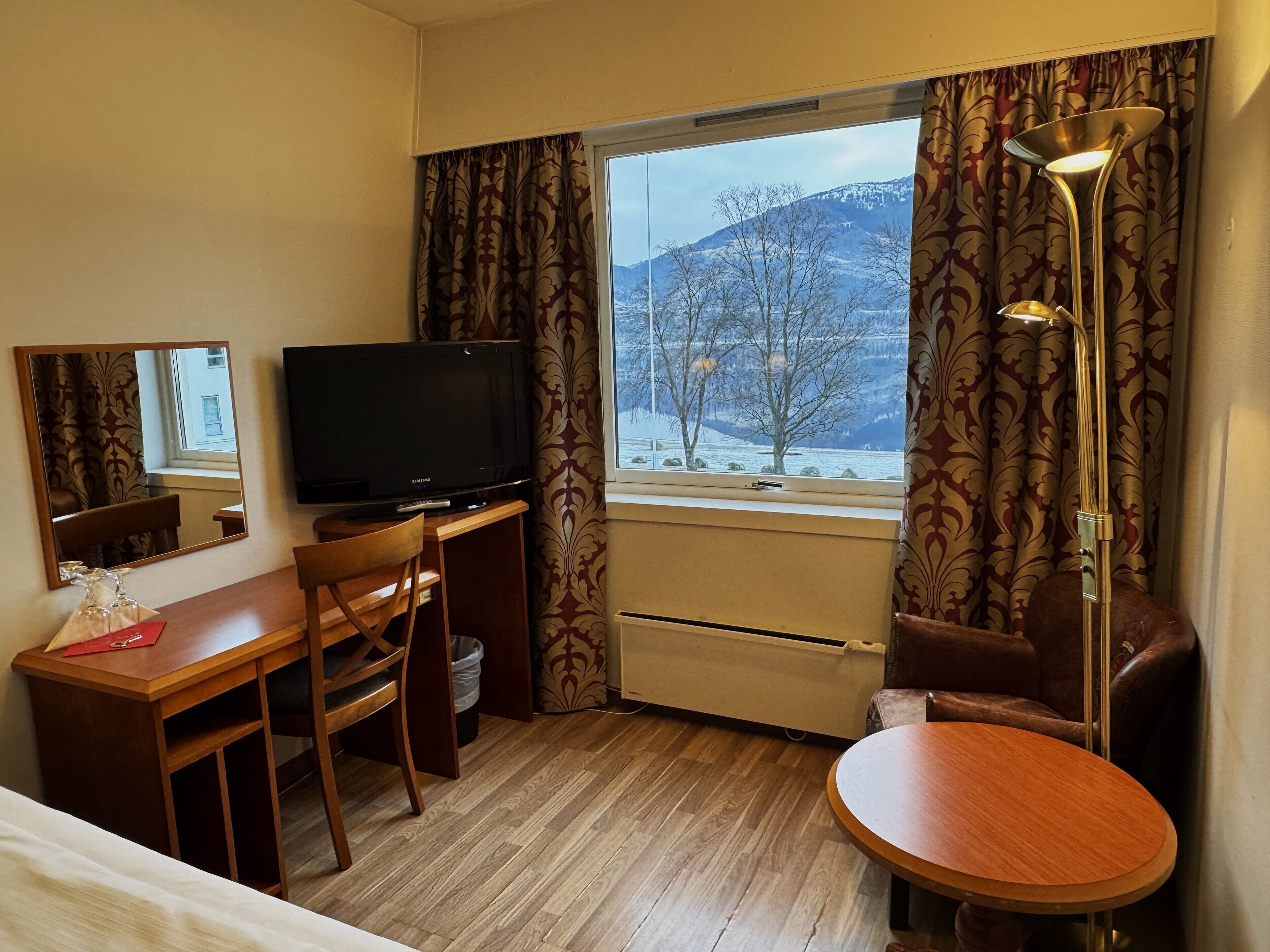 A hotel room with a wooden desk, a television, a chair, a small table, a mirror, and a window with a view of snow-covered mountains.