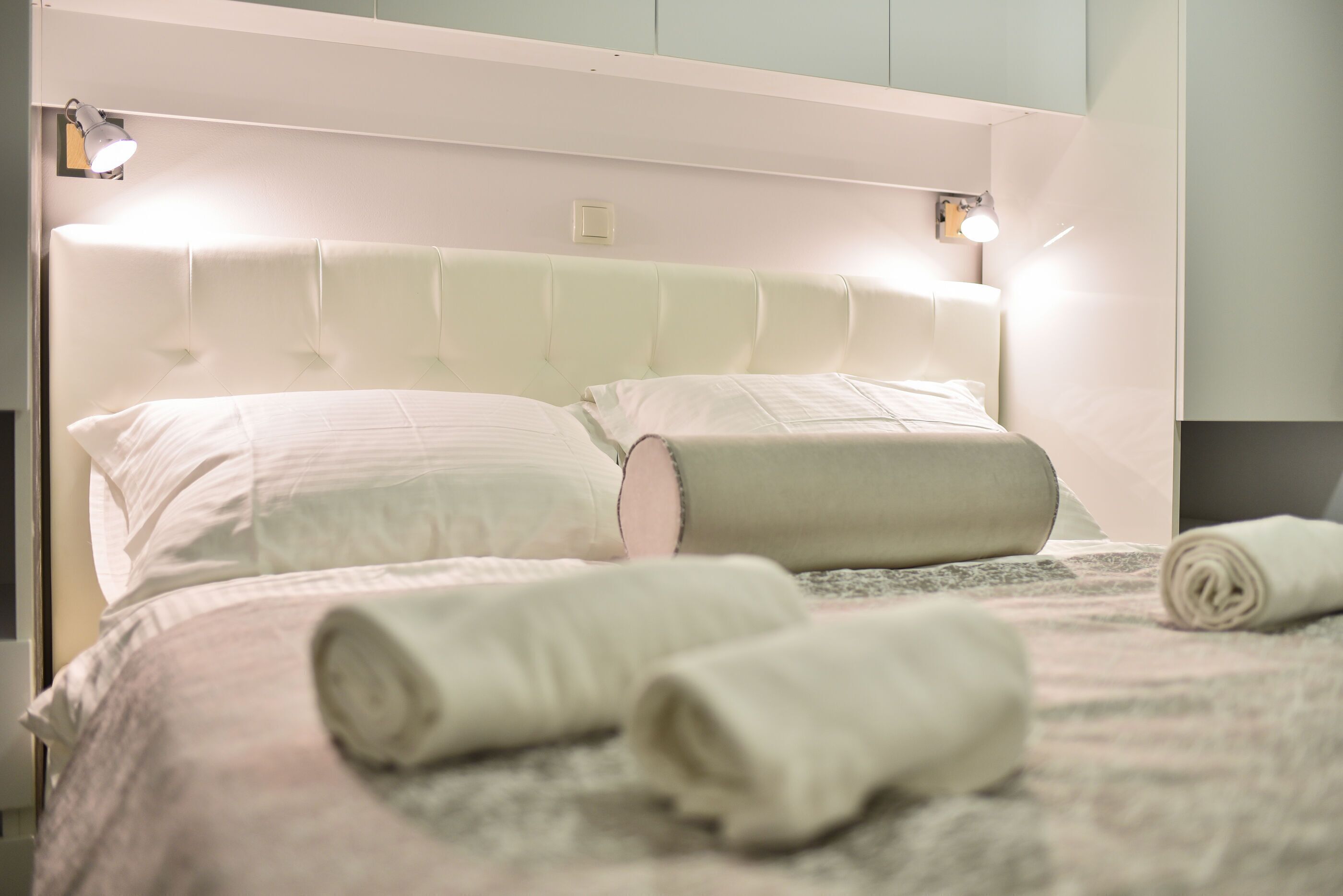 Una cama bien hecha con toallas enrolladas, un cabecero capitonado y luces de lectura fijadas a la pared.