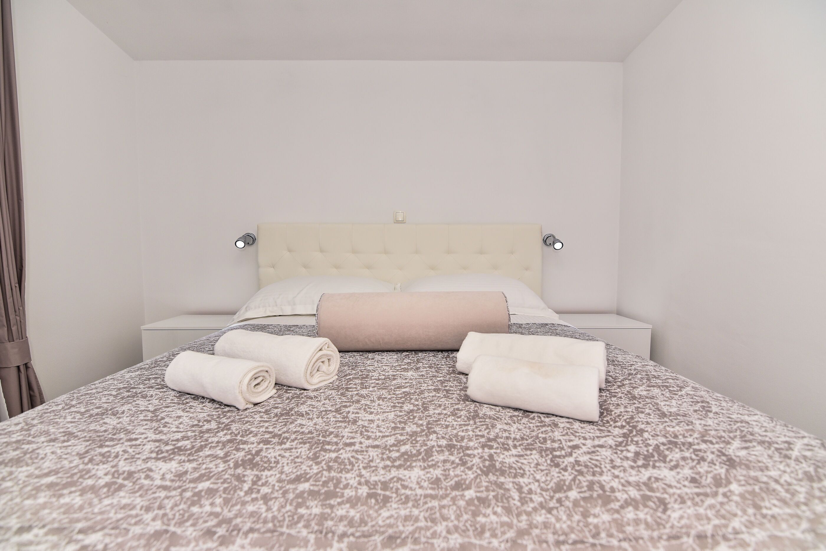 A neatly made bed with a patterned bedspread and rolled towels, a tufted headboard, and a bedside table with a lamp.