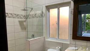 A bathroom with a glass-enclosed shower, a toilet, a sink with toiletries, and a window with a view of greenery. - Come have Fun in DR! (Sosúa)