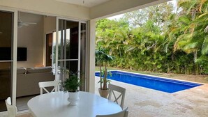 A patio area with a round table, chairs, a ceiling fan, and a view of a swimming pool surrounded by lush greenery. - Come have Fun in DR! (Sosúa)