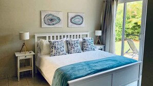 A bedroom with a white bed, two wall art pieces, a nightstand with a lamp, and a view of greenery through a window. - Come have Fun in DR! (Sosúa)